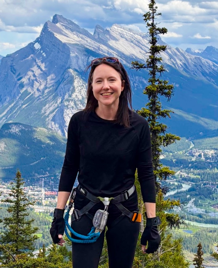 Michelle Walstra on the Mount Norquay via ferrata above Banff, Alberta — climbing harness on, Mt. Rundle behind her